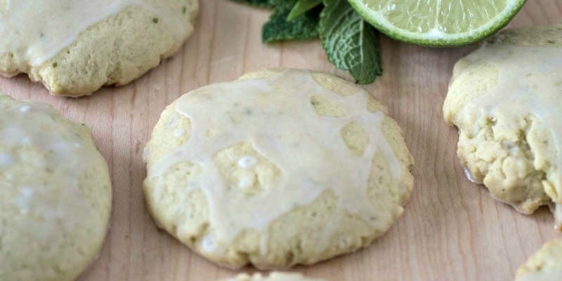 Mojito Sugar Cookies with a white glaze sit on a wooden surface, accompanied by a halved lime and fresh mint leaves.