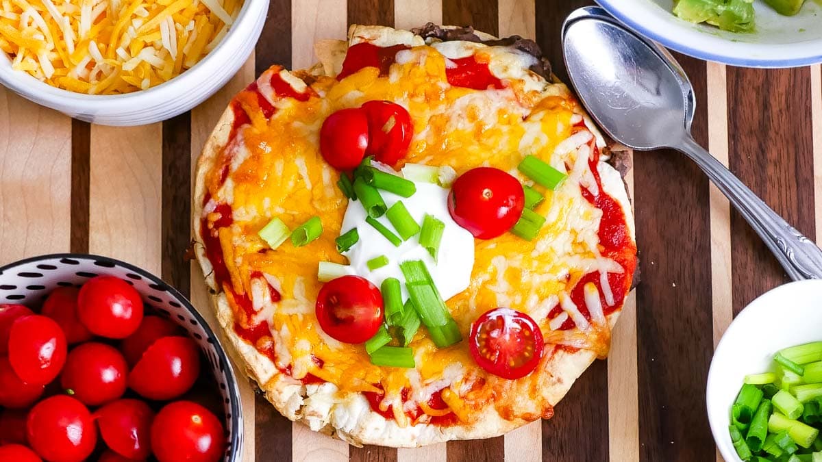 A homemade pizza inspired by Taco Bell's Mexican Pizza, topped with cheese, tomatoes, and green onions, surrounded by bowls of ingredients and a spoon on a wooden table.
