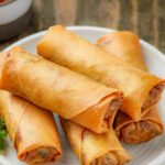 A plate with six golden-brown spring rolls stacked on it. Text reads "Air Fryer Spring Rolls" and "Game Day Favorite" with a football icon—perfectly crispy and delicious for any sports gathering.