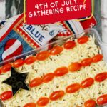 An image of a bean dip decorated like the american flag with tomatoes, cheese, and a black olive, labeled as a "4th of july gathering recipe.