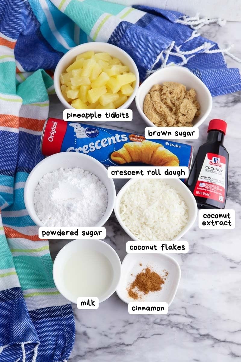 Ingredients for baking displayed on a marble surface, including crescent roll dough, brown sugar, powdered sugar, milk, coconut flakes, coconut extract, cinnamon, and pineapple tidbits.
