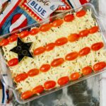 A bean dish with shredded cheese topping and cherry tomatoes arranged in stripes, mimicking the american flag, on a patriotic-themed napkin.
