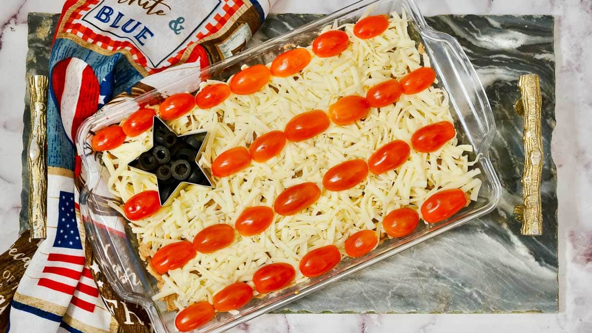 A bean dish with shredded cheese topping and cherry tomatoes arranged in stripes, mimicking the american flag, on a patriotic-themed napkin.