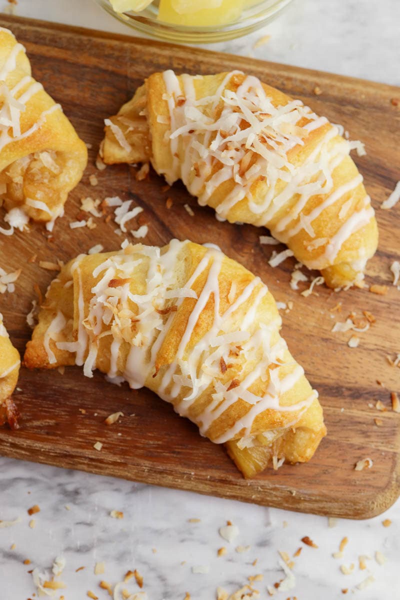 Freshly baked pineapple crescent roll topped with glaze and shredded coconut, served on a wooden cutting board.