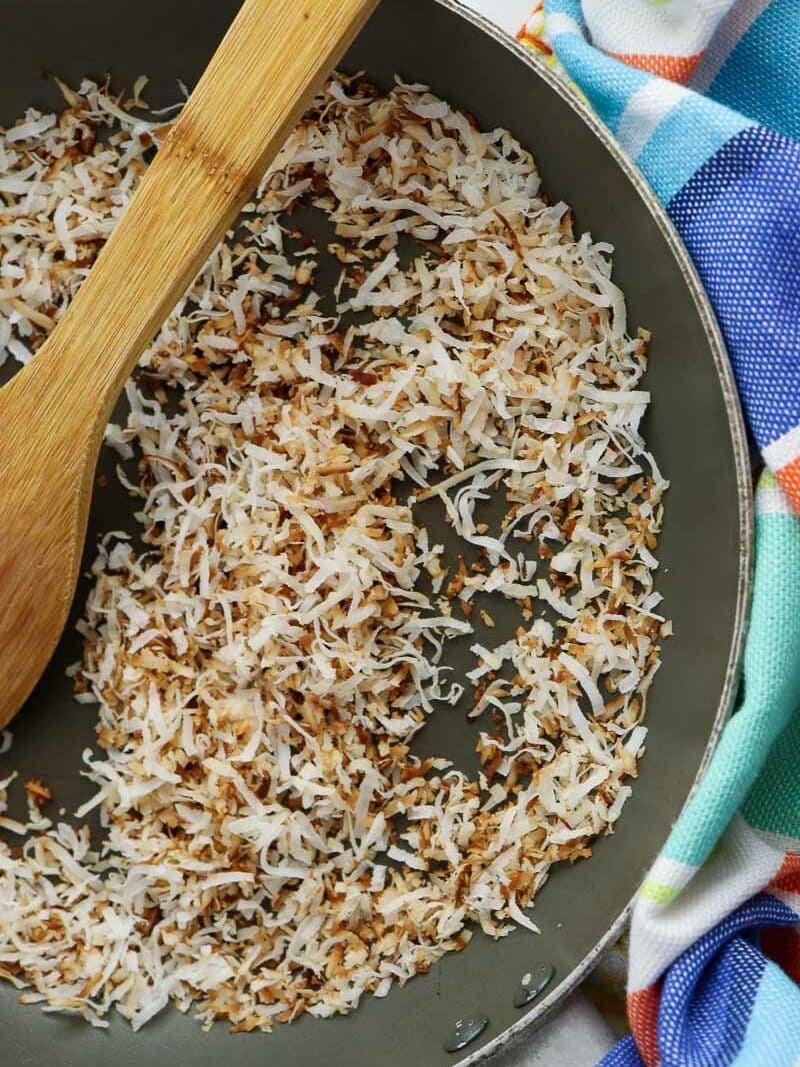 Fried shredded coconut in a skillet with a wooden spoon, beside a colorful cloth.