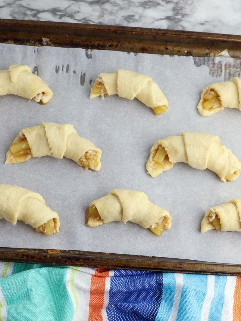 Unbaked crescent roll with pineapple filling on a parchment-lined baking sheet, with a colorful striped towel on the side.
