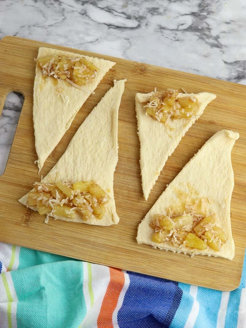 Four slices of crescent roll dough topped with pineapple mixture on a wooden cutting board, with a colorful striped towel in the background.