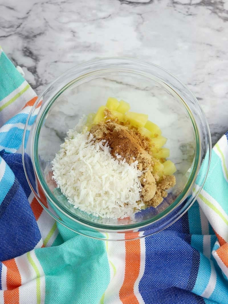 A bowl of ingredients for baking on a marble countertop, including pineapple, brown sugar, cinnamon, and 1/4 cup coconut flakes.