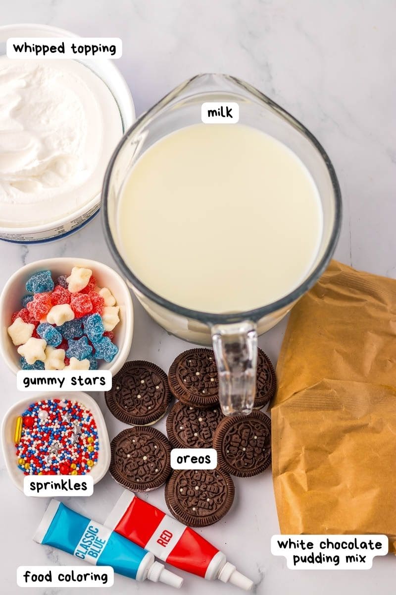 A variety of dessert ingredients on a counter including whipped topping, a jug of milk, white chocolate pudding mix, Oreos, sprinkles, gummy stars, and tubes of blue and red food coloring.