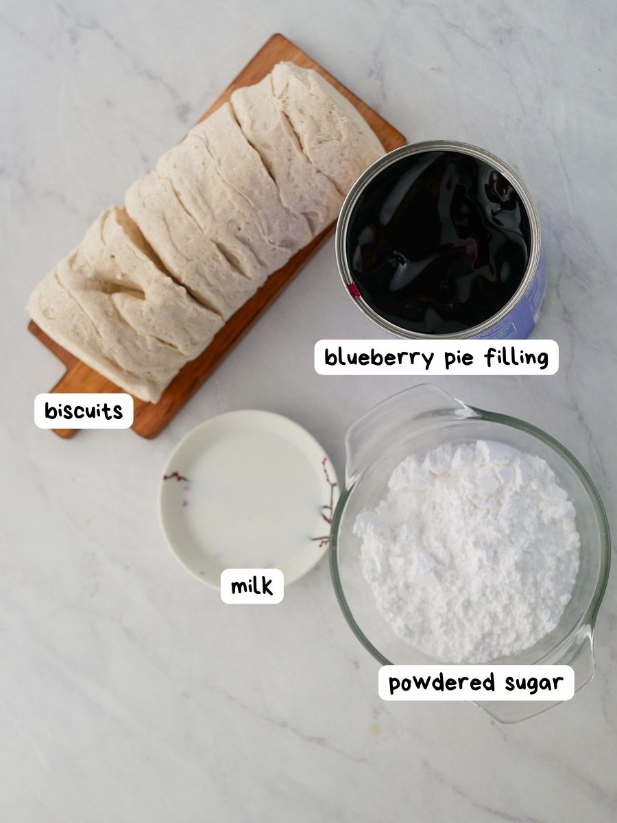 Top-down view of baking ingredients including sliced biscuits, a can of blueberry pie filling, a cup of powdered sugar, and a small bowl of milk, each labeled accordingly.