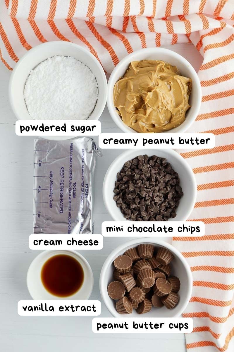 Ingredients for a recipe: powdered sugar, creamy peanut butter, mini chocolate chips, cream cheese, vanilla extract, and peanut butter cups, arranged in bowls on a striped cloth.