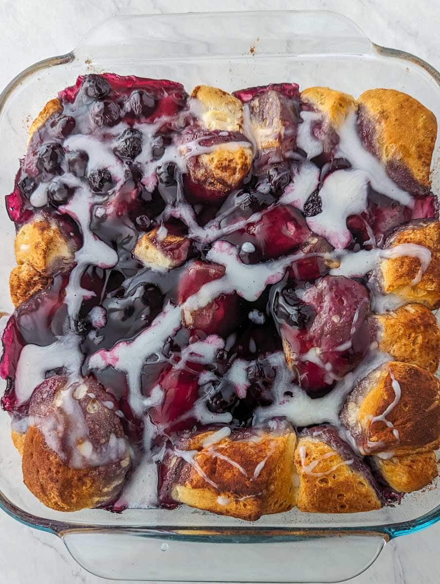 A glass baking dish contains a baked dessert with golden-brown dough pieces, Blueberry Pie Filling, and a white drizzle topping.