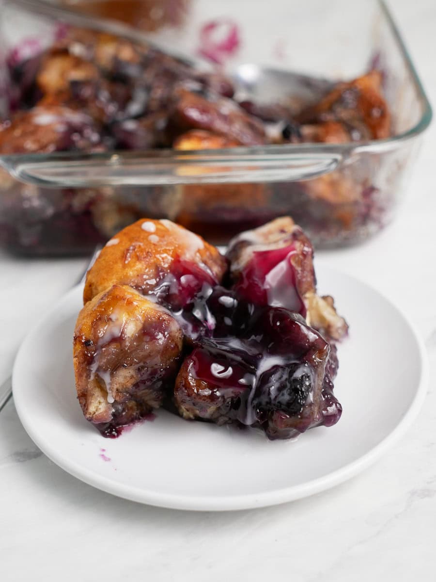 A white plate with a serving of warm, glazed Blueberry Bubble Up in front of a glass baking dish containing more cobbler.