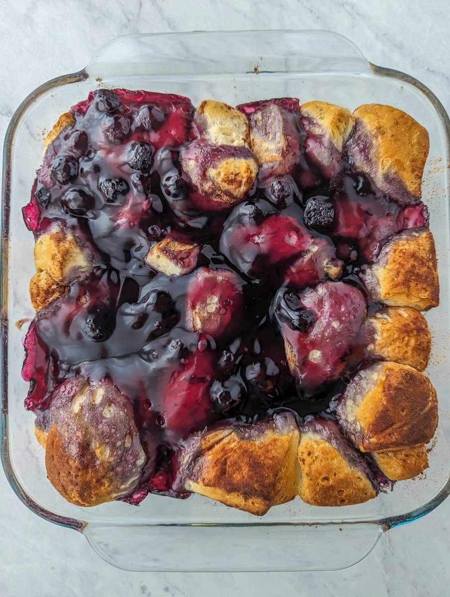 A glass baking dish filled with golden-brown pull-apart bread topped with dark berry jam.