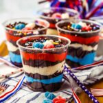 Clear plastic cups filled with layered desserts in red, white, and blue colors, topped with sprinkles and gummy candy, are displayed on an American-themed table setting.