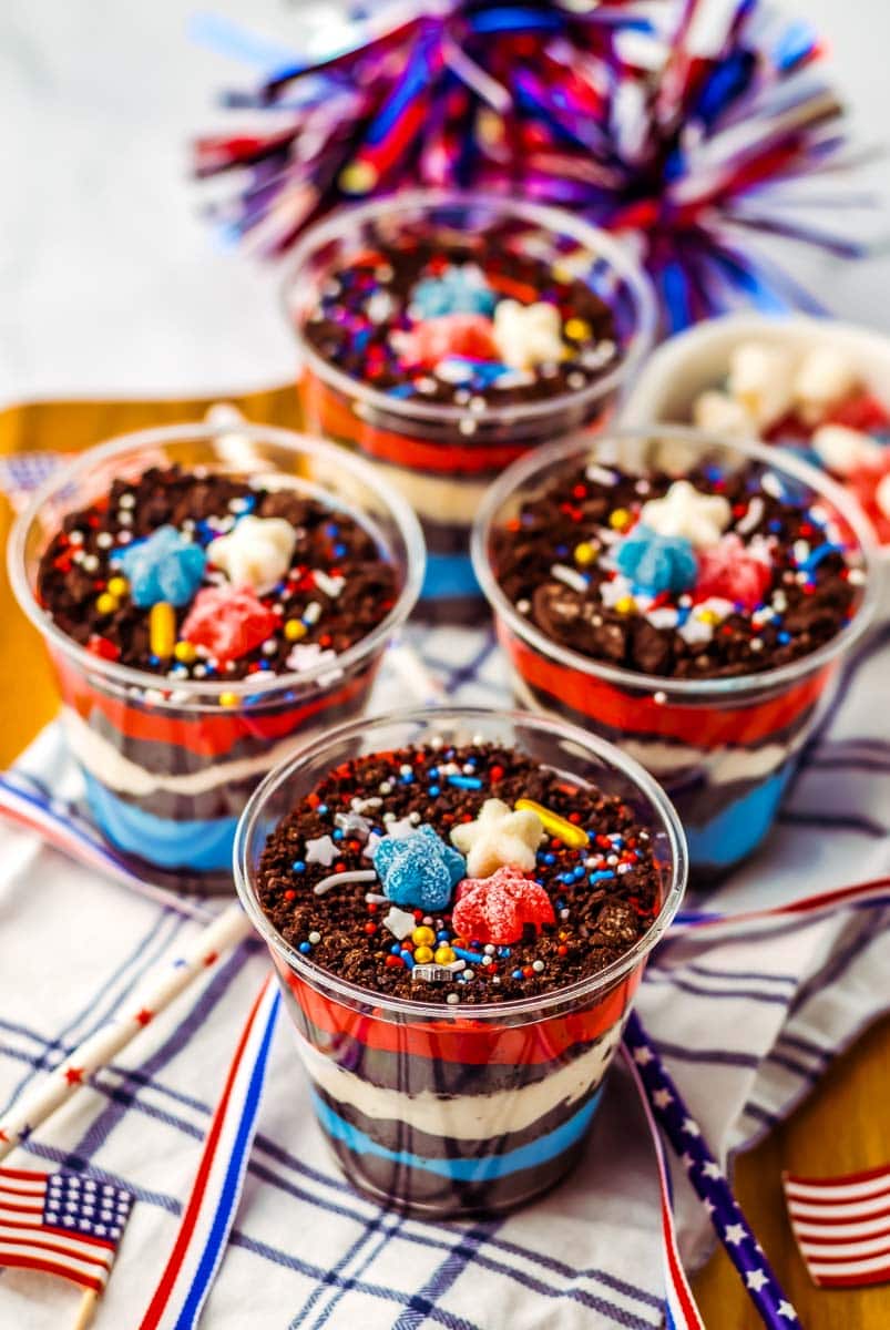 Four cups of layered dessert with red, white, and blue colors, topped with chocolate crumbs and star-shaped candies. American flags and patriotic decorations are placed around the cups.