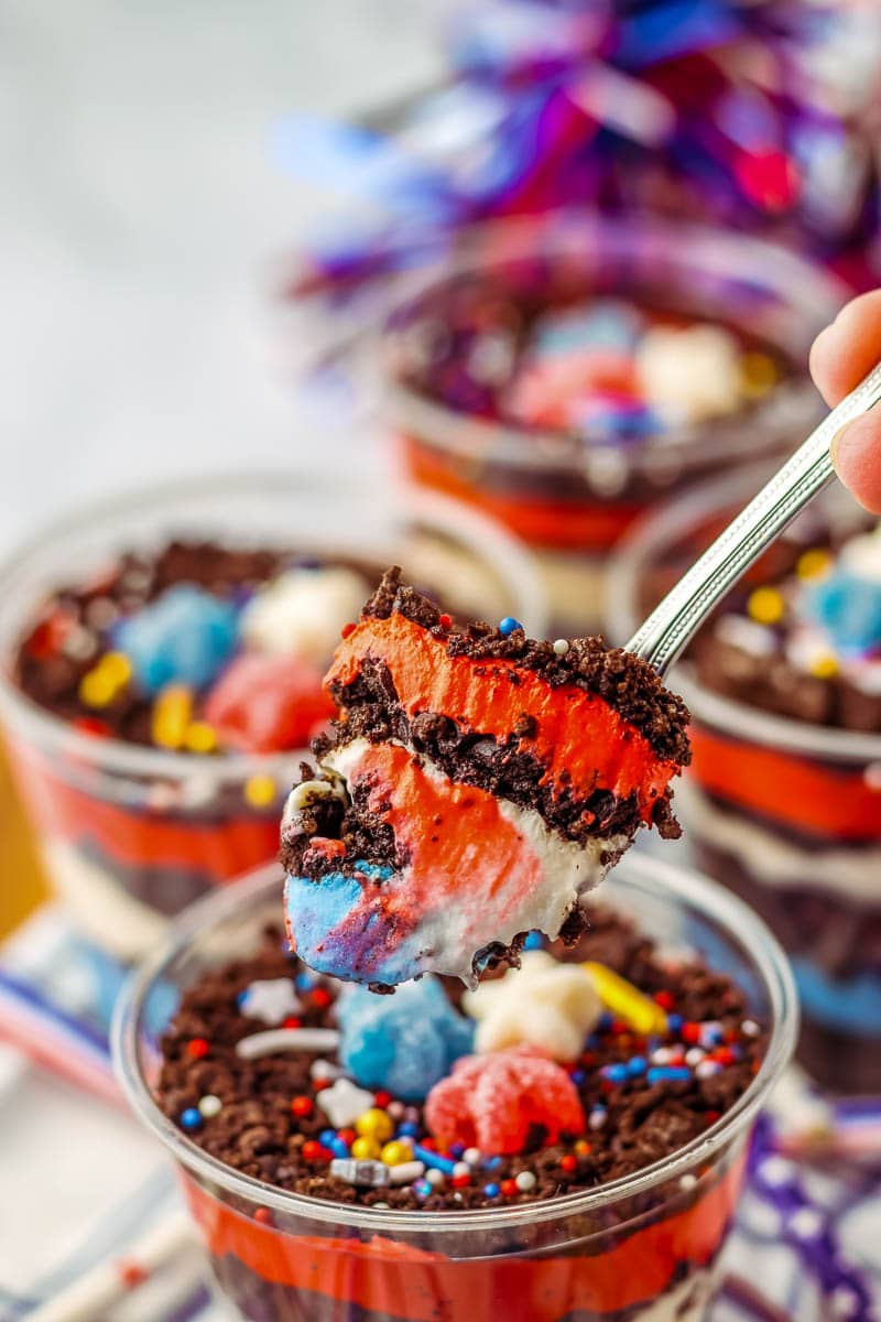 A spoonful of layered dessert featuring red, white, and blue colors along with Oreo crumbles and colorful sprinkles. More servings of the dessert are visible in the background.