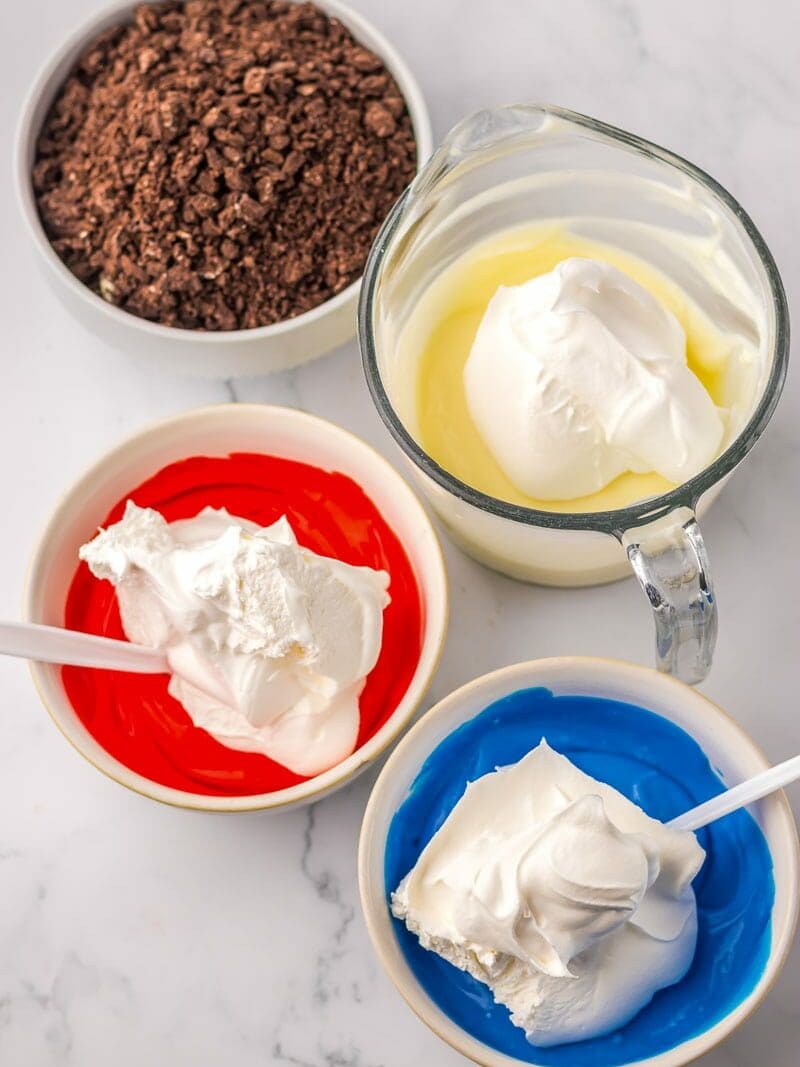 Three small bowls containing whipped topping mixed with red, blue, and yellow ingredients are on a marble surface. There is also a bowl of chocolate crumbles in the background.