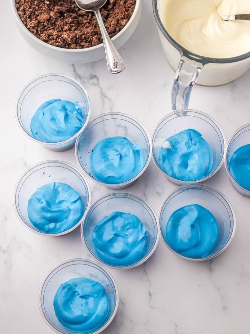 Eight small plastic cups filled with blue whipped cream, a bowl of crushed chocolate with a spoon, and a measuring cup containing white whipped cream on a marble surface.