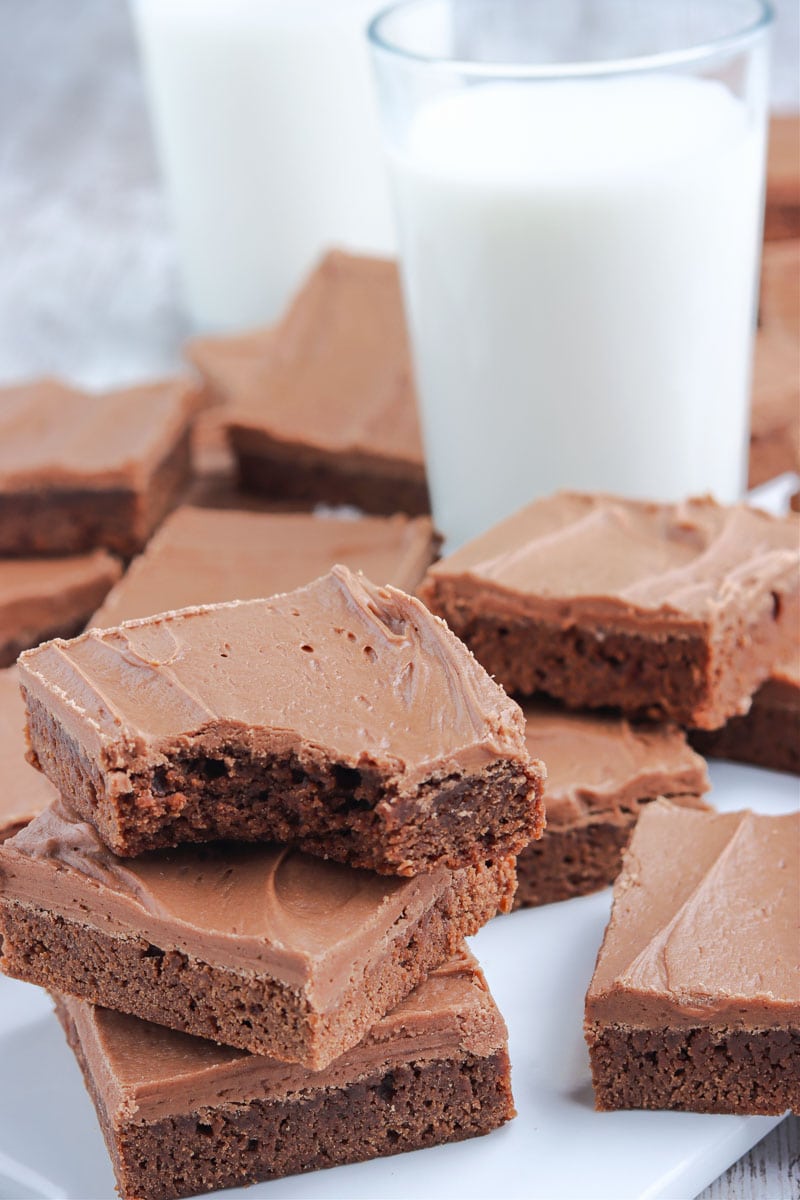 A stack of chocolate frosted brownies with a bite taken out of the top brownie, placed next to two glasses of milk.