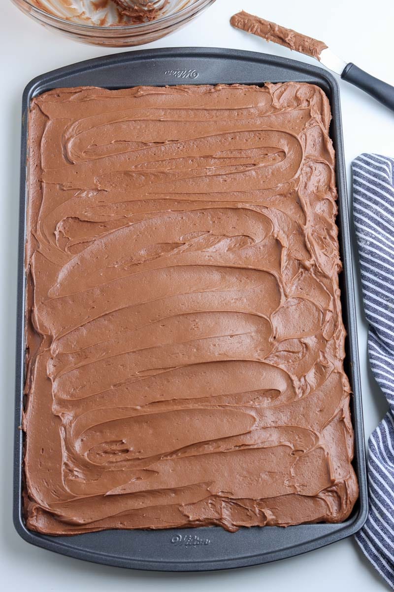 A baking sheet with chocolate frosting spread evenly over the surface. A spatula with some chocolate frosting and a striped cloth are beside the sheet.