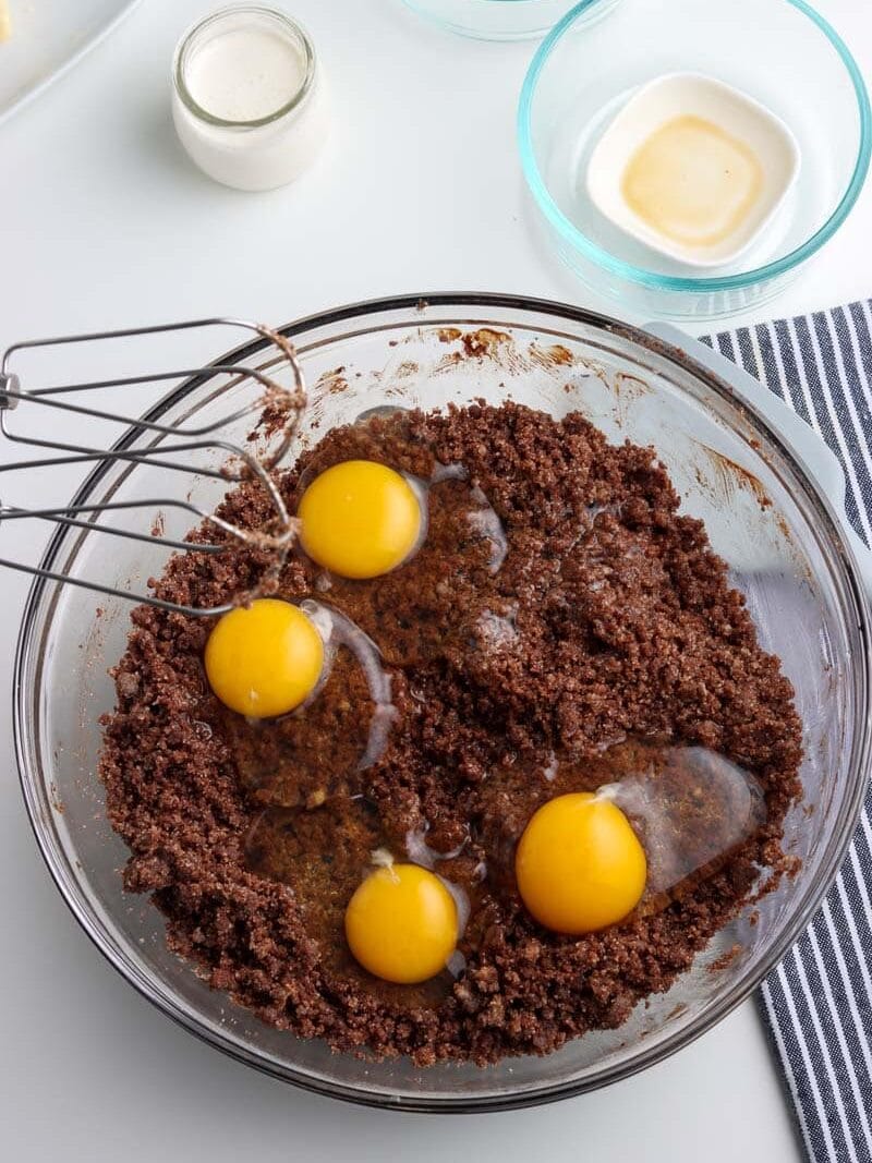 A mixing bowl with crumbled chocolate mixture and four cracked egg yolks. A whisk attachment, a small jug of milk, and empty bowls are placed around it on a white surface with a striped cloth.