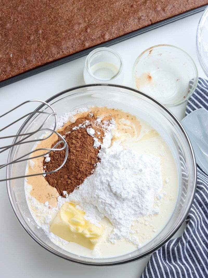 A mixing bowl with ingredients for frosting, including butter, powdered sugar, cocoa powder, and liquid, with a whisk attachment and brown baked cake in the background.