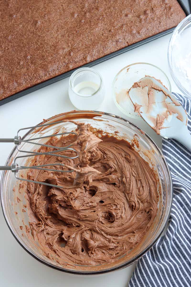 A glass bowl filled with chocolate frosting being mixed with an electric mixer. Surrounding the bowl are a large slab of baked cake, a small container of white frosting, and a striped cloth.