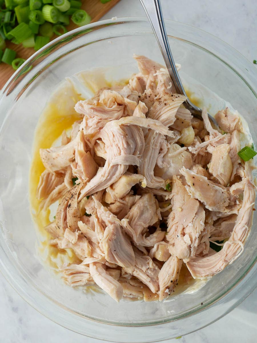 Shredded chicken in a glass bowl with a fork, seasoned with herbs and surrounded by sliced green onions.