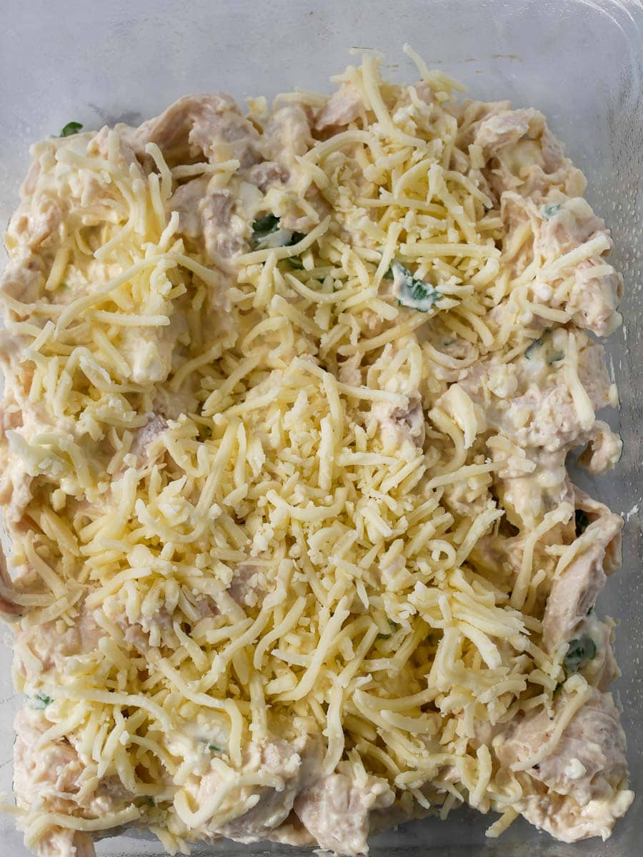 A glass baking dish filled with uncooked creamy chicken pasta topped with shredded cheese, ready for baking.