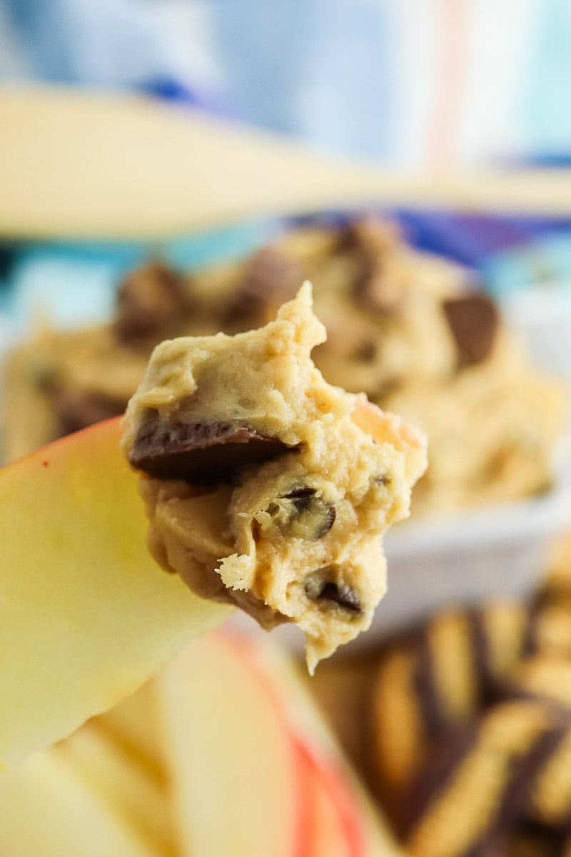 A hand holds a slice of apple dipped in peanut butter dip with chocolate chunks.