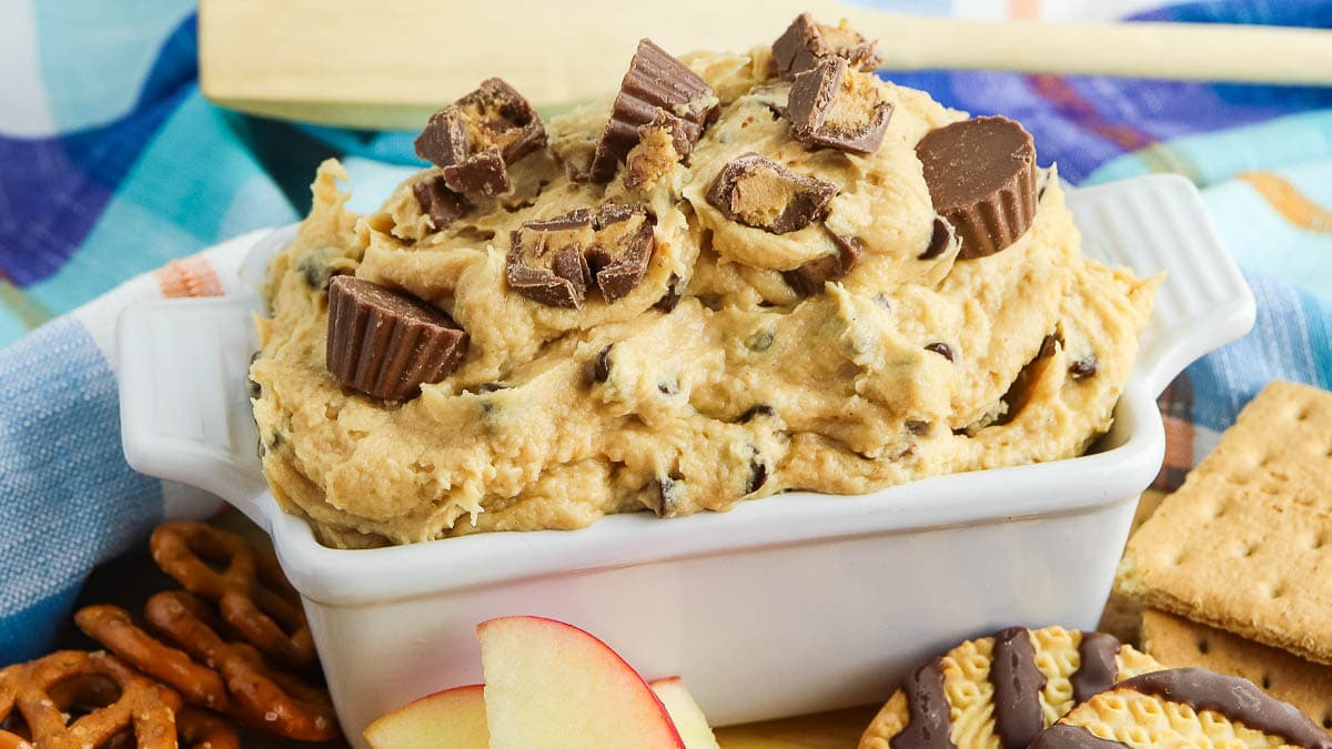 A dish of peanut butter dip topped with chocolate pieces served with apple slices and pretzels on the side.