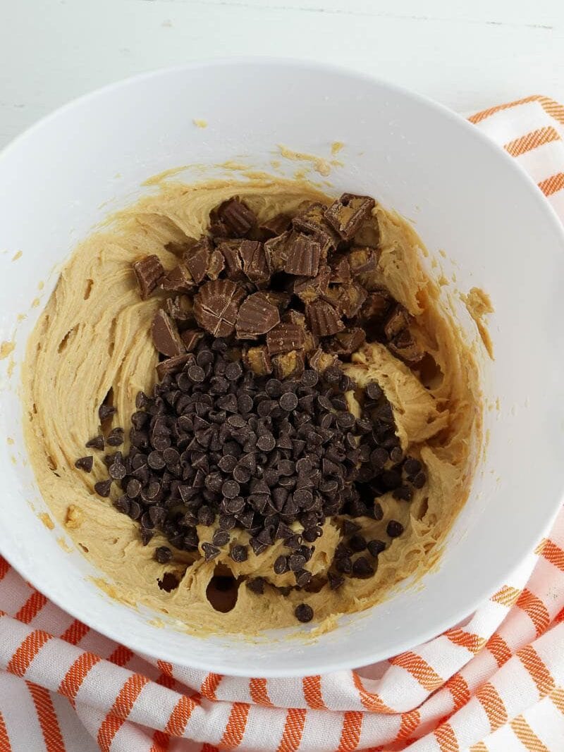 A white bowl filled with peanut butter, chocolate chips, and chopped chocolate pieces on a white and orange striped cloth.