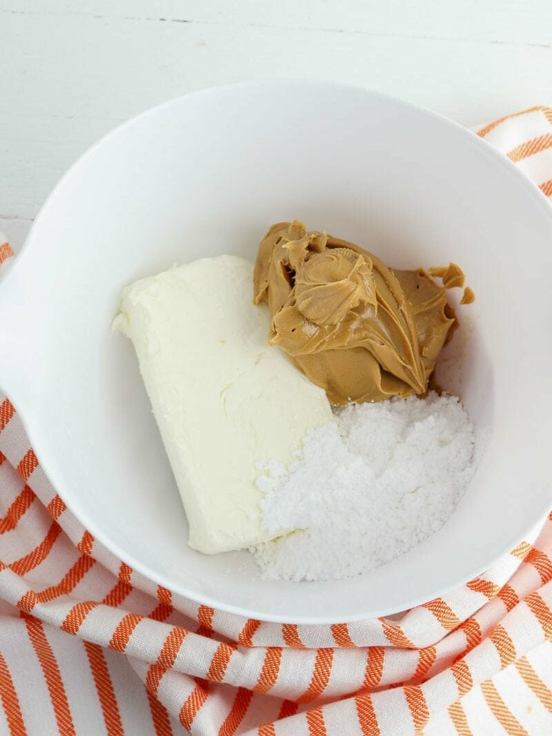A white bowl contains cream cheese, peanut butter, and powdered sugar. It is placed on a white and orange striped cloth.