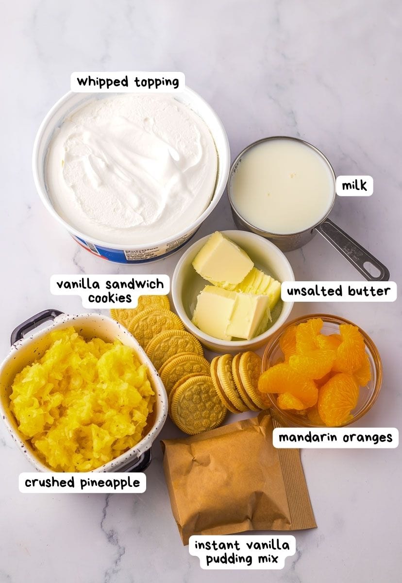 Ingredients on a counter: whipped topping, milk, vanilla sandwich cookies, unsalted butter, mandarin oranges, crushed pineapple, and instant vanilla pudding mix.
