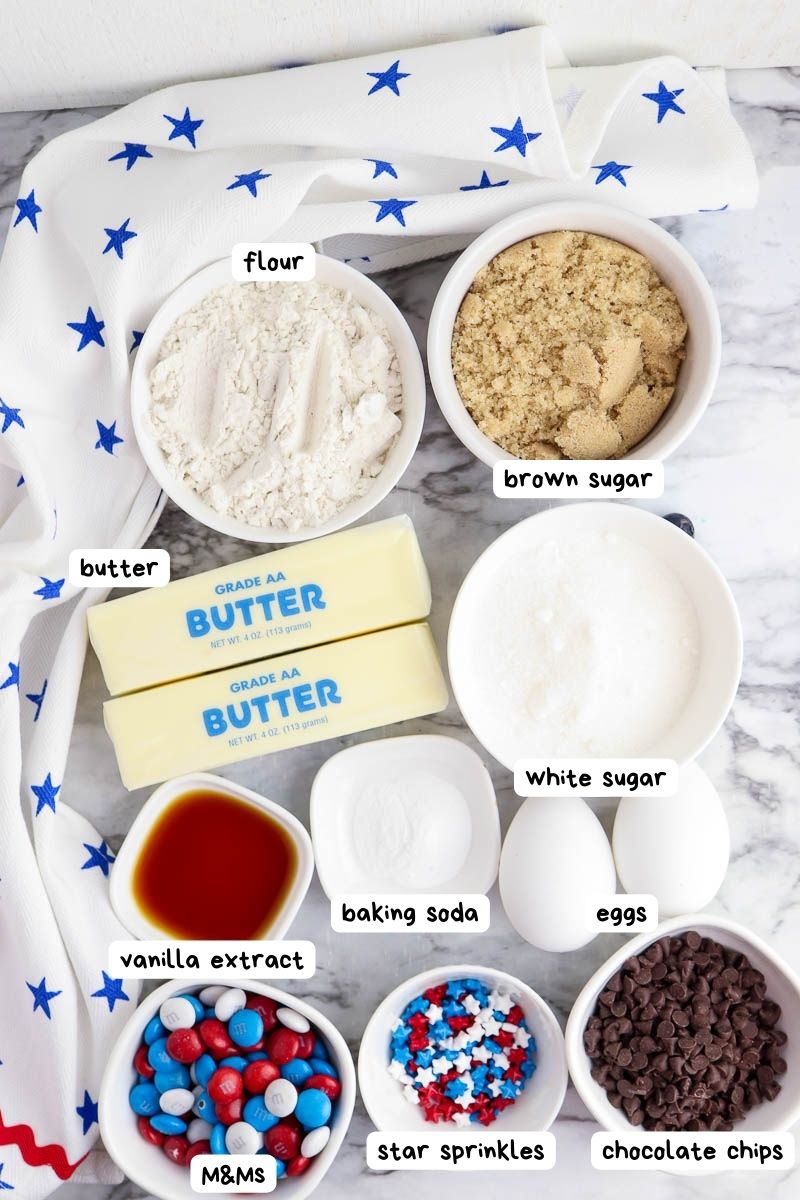 Ingredients for baking displayed: flour, brown sugar, butter, white sugar, vanilla extract, baking soda, eggs, M&Ms, star sprinkles, and chocolate chips. Background includes a white cloth with blue stars.
