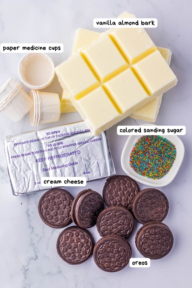 An assortment of ingredients including vanilla almond bark, cream cheese, Oreos, colored sanding sugar, and paper medicine cups is arranged on a marble surface.