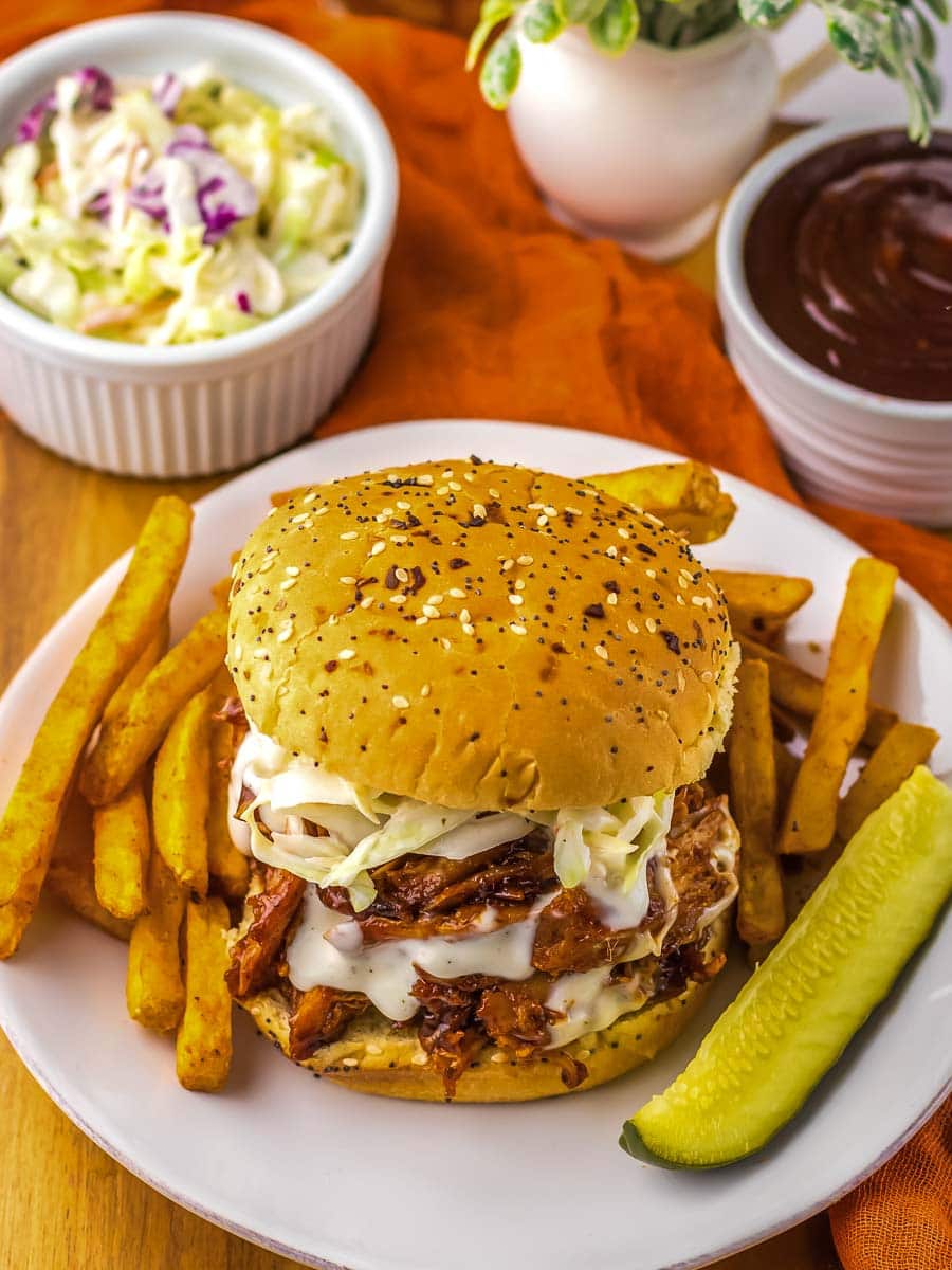 A plate with a pulled pork burger topped with coleslaw, accompanied by French fries and a pickle slice. In the background are side dishes of coleslaw and barbecue sauce.
