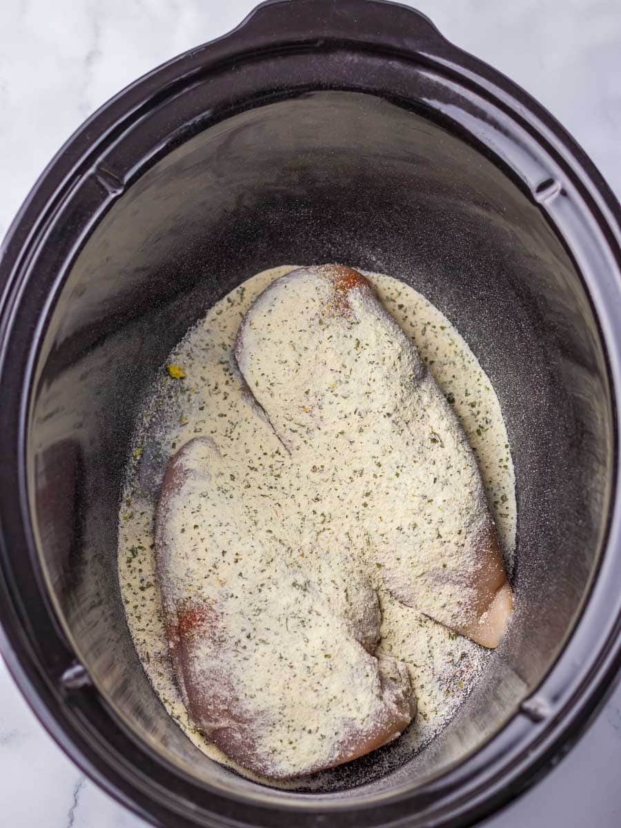 Two seasoned raw chicken breasts in a black slow cooker pot on a marble surface.