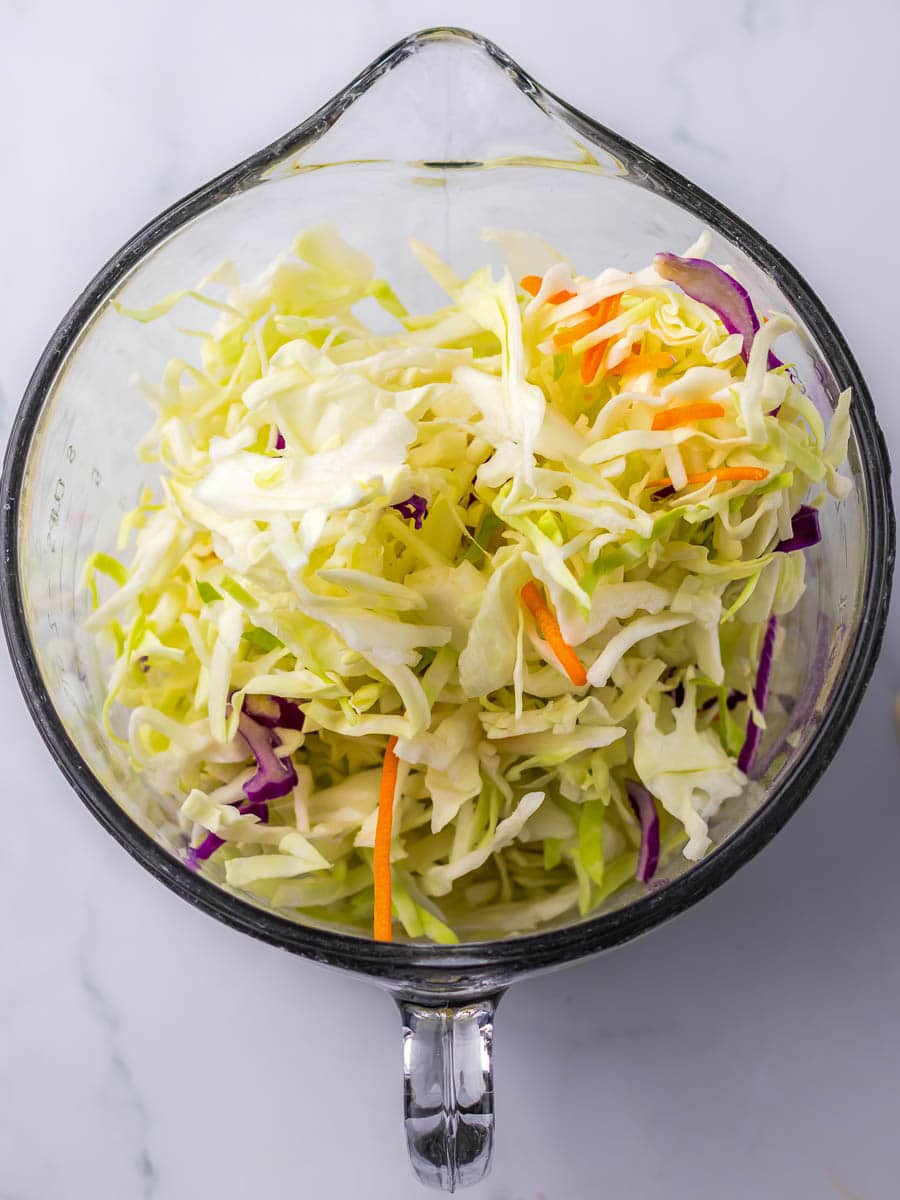 A clear glass bowl filled with shredded cabbage, including green, purple, and orange pieces, atop a white surface.