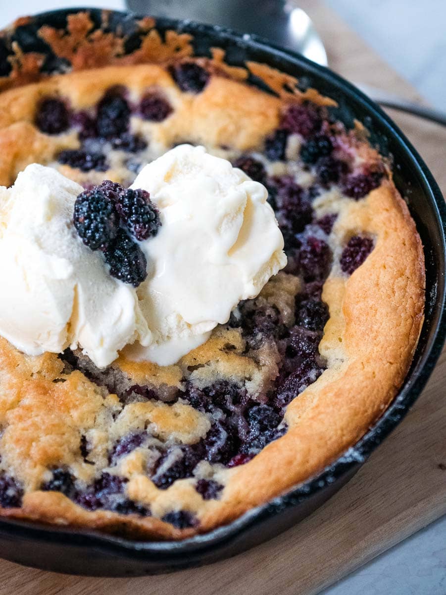 A freshly baked blackberry cobbler in a black pan, topped with a scoop of vanilla ice cream and garnished with blackberries.