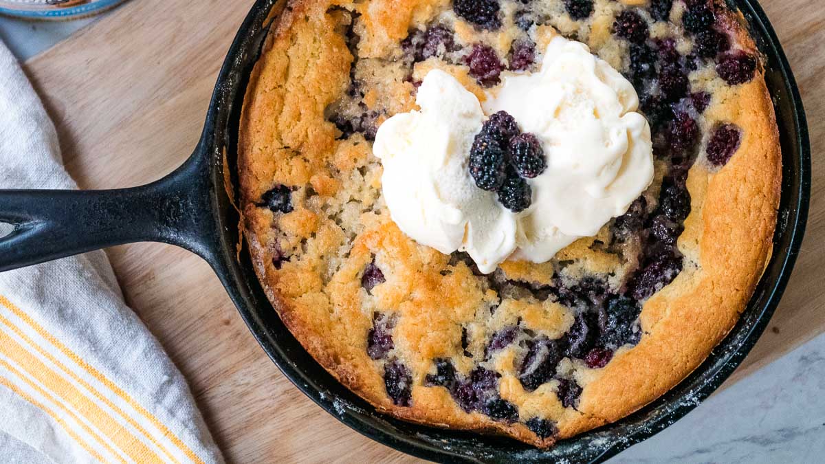 Cast iron skillet with a freshly baked blackberry cobbler topped with a scoop of vanilla ice cream and extra blackberries on a wooden surface next to a white dish towel with yellow stripes.