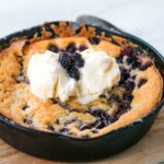 A cast iron skillet with a baked berry cobbler topped with a scoop of vanilla ice cream and garnished with fresh blackberries.