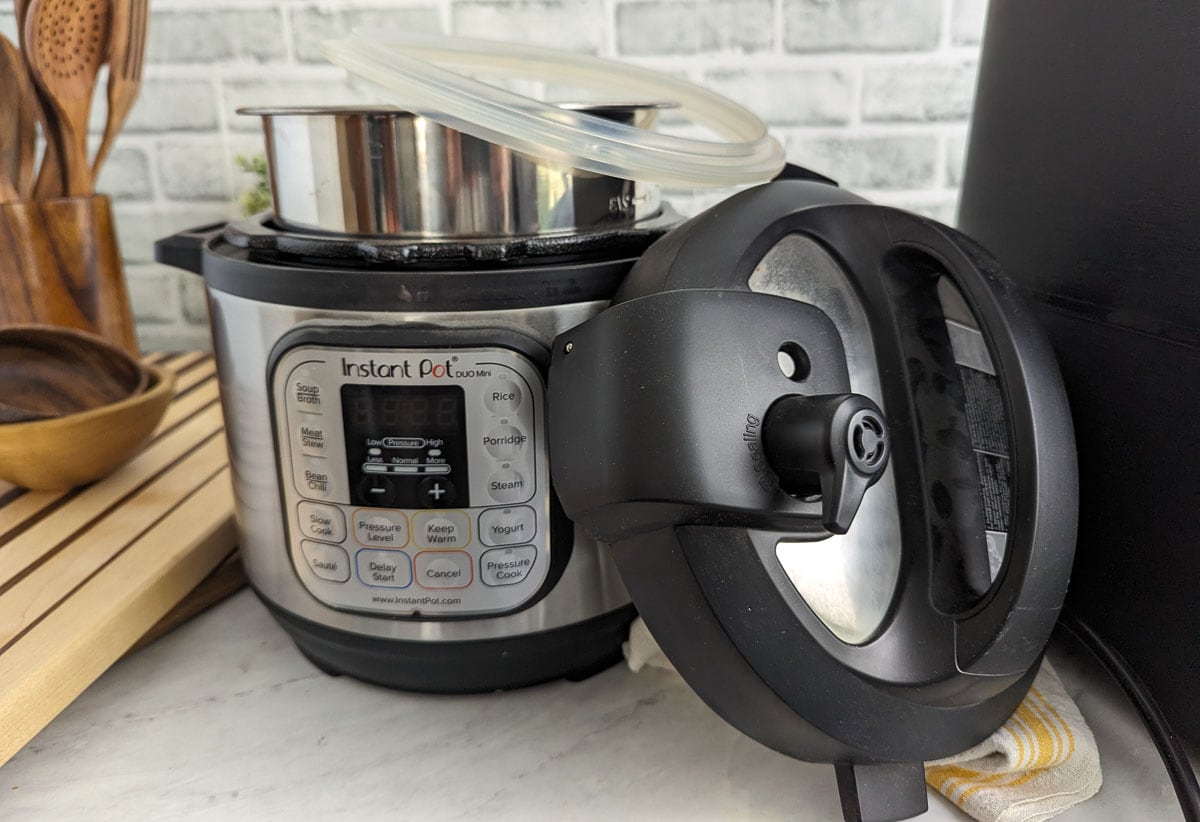An Instant Pot stands on a kitchen counter with its lid propped open. A silicone sealing ring and a metal pot are placed inside. The control panel displays various cooking options.