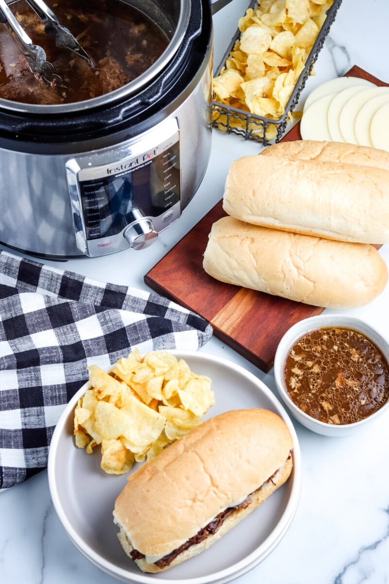 A plate with a sandwich and potato chips is placed near a slow cooker with meat, a bowl of sauce, uncut sandwich rolls, sliced cheese, and a basket of chips on a marble surface.