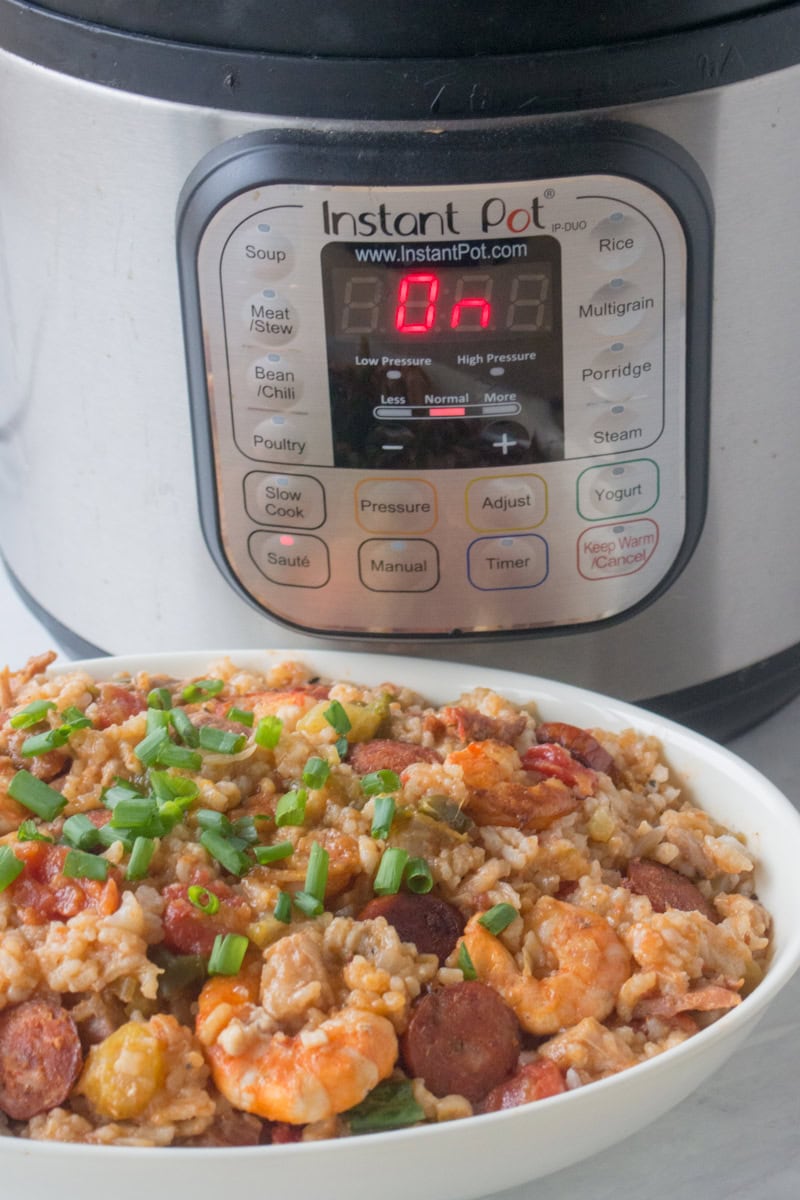 A bowl of shrimp and sausage jambalaya garnished with chopped green onions is placed in front of an Instant Pot. The Instant Pot display shows "L0:00.
