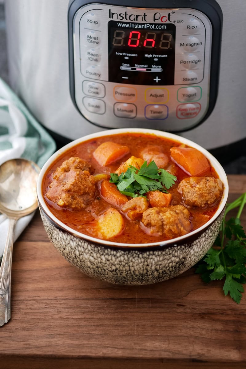 A bowl of stew with meatballs, potatoes, and carrots sits in front of an Instant Pot displaying "B-A." A spoon and a sprig of parsley are placed nearby.