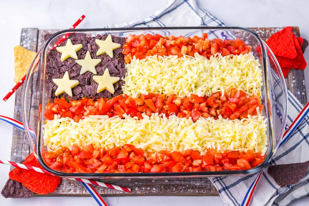 A layered dip arranged to look like the American flag, made with blue tortilla chips, cheese stars, shredded cheese, and diced tomatoes. Two American flag-themed napkins are placed nearby.