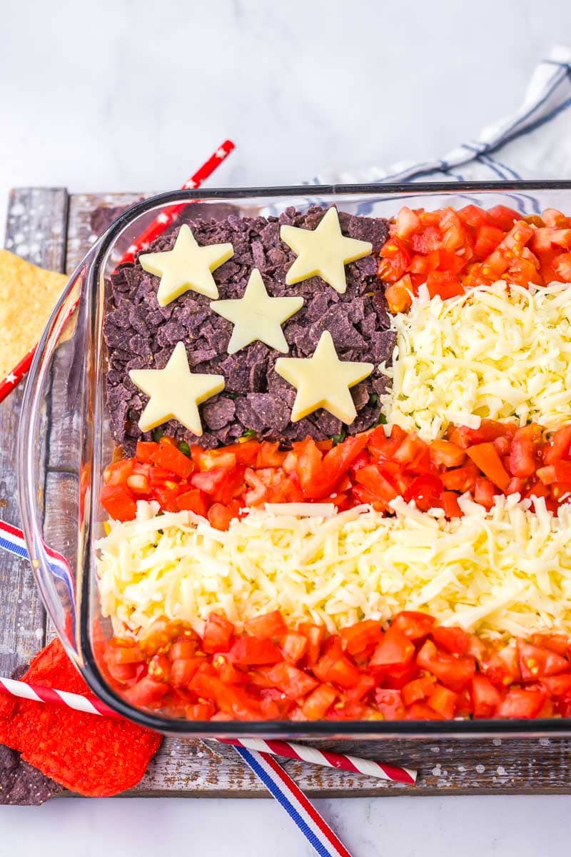A layered dip in a glass dish, with tortilla chips, diced tomatoes, and shredded cheese arranged to resemble the American flag.