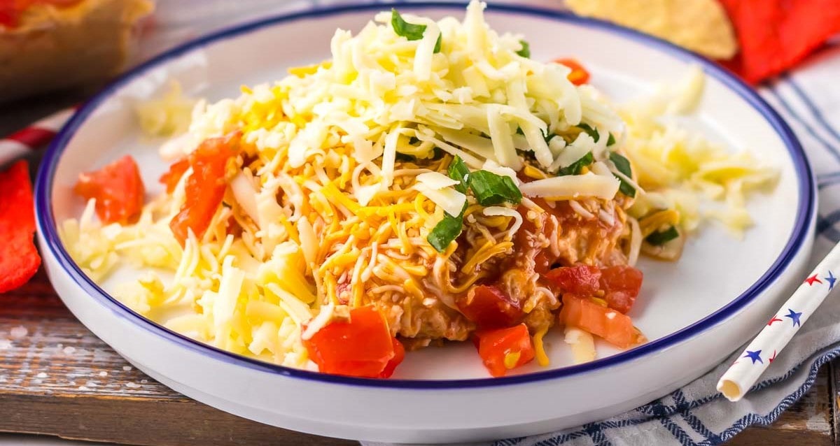 A plated dish of layered taco dip with shredded cheese, diced tomatoes, lettuce, and ground meat, elegantly displayed on a white plate with a blue rim.